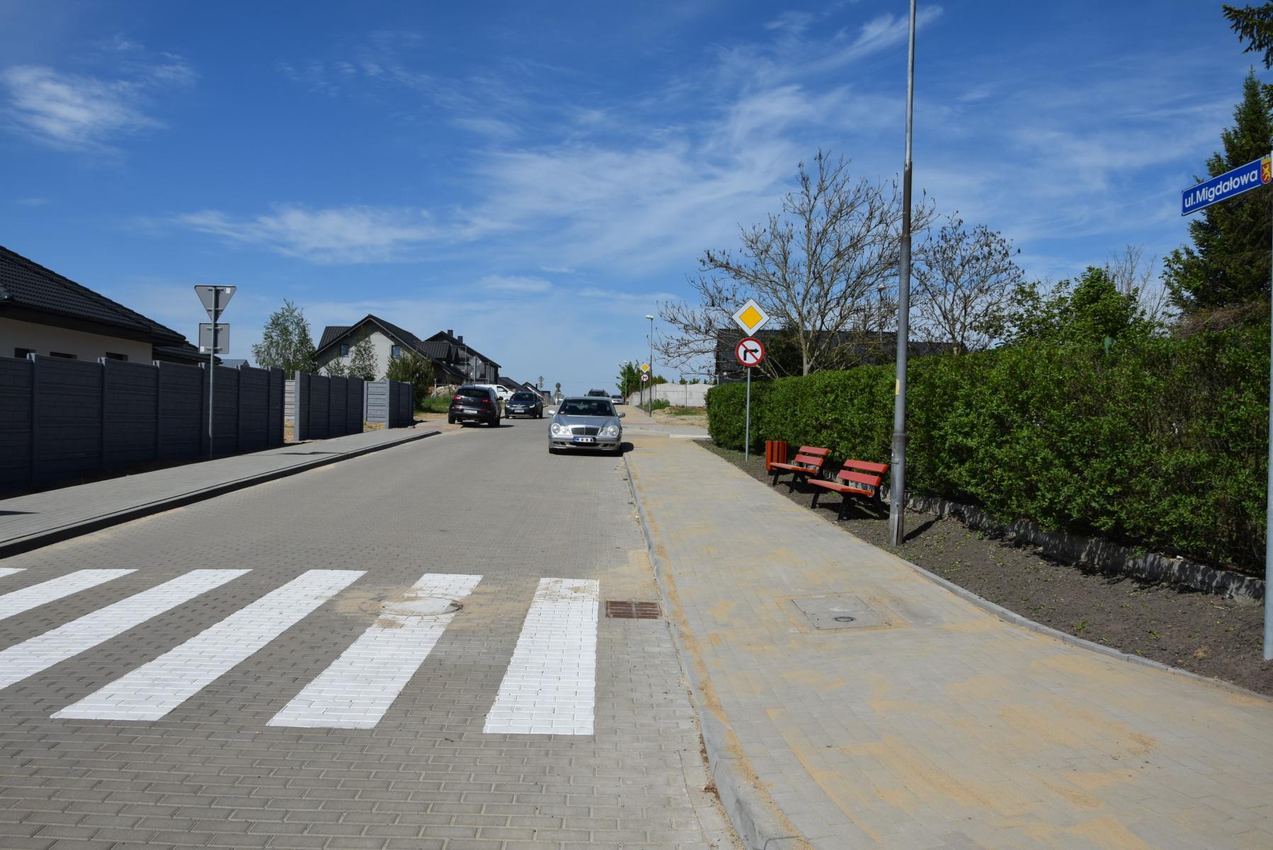 Ulica Migdałowa, z prawej stoją ławeczki i kosz na śmieci, znak zakaz skrętu w prawo. samochody na jezdni, pasy na jezdni