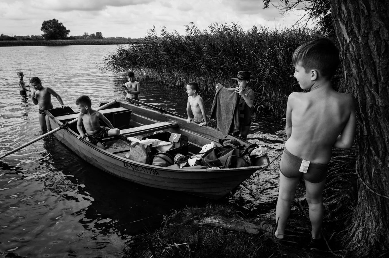 Czarno-biała fotografia. Chłopcy na brzegu jeziora stoją, jeden siedzi w łódce