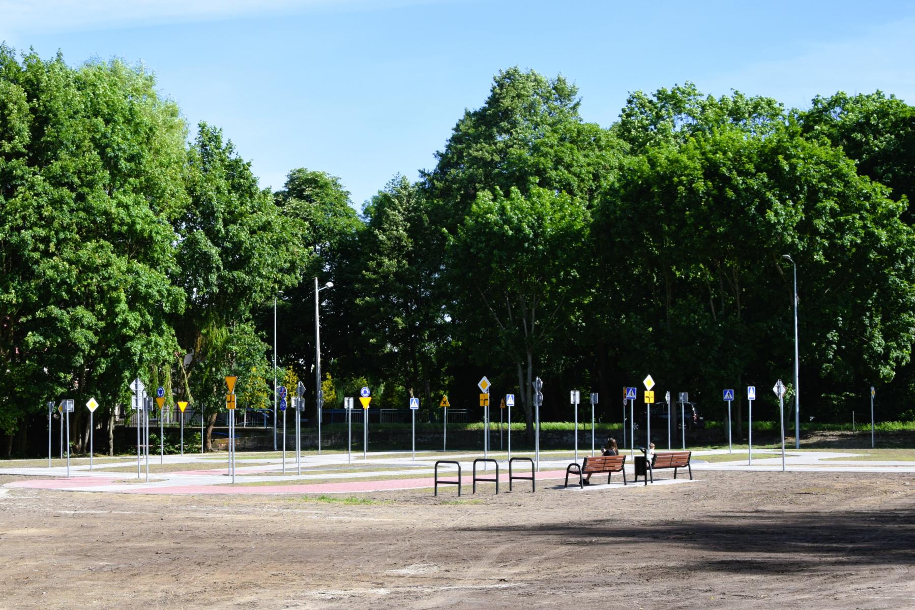 Cały plac z miasteczkiem rowerowym. W tle duże zielone drzewa, niebieskie niebo