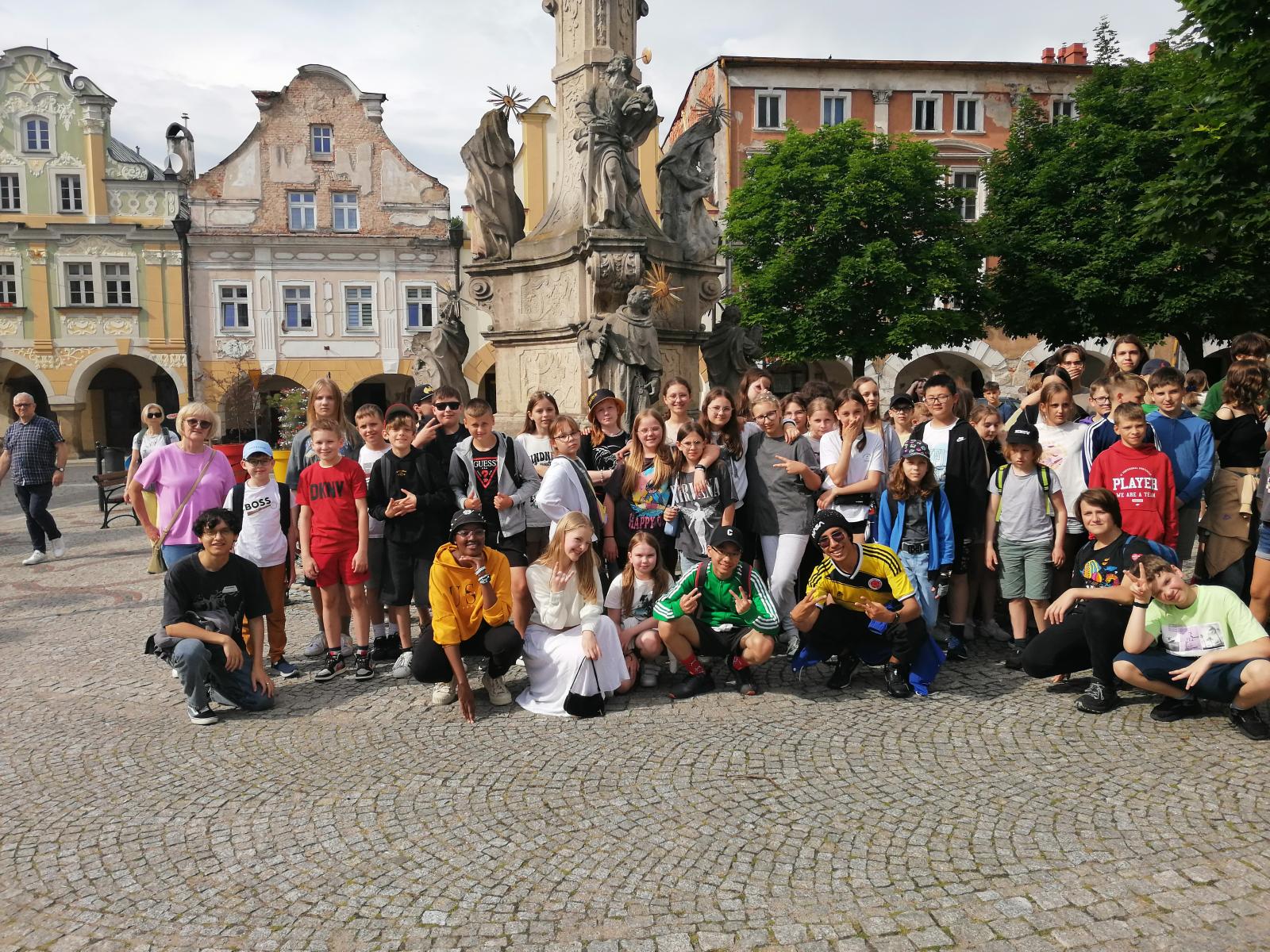 Grupa dzieci, stoją na placu, z tyłu kamienice i pomnik 