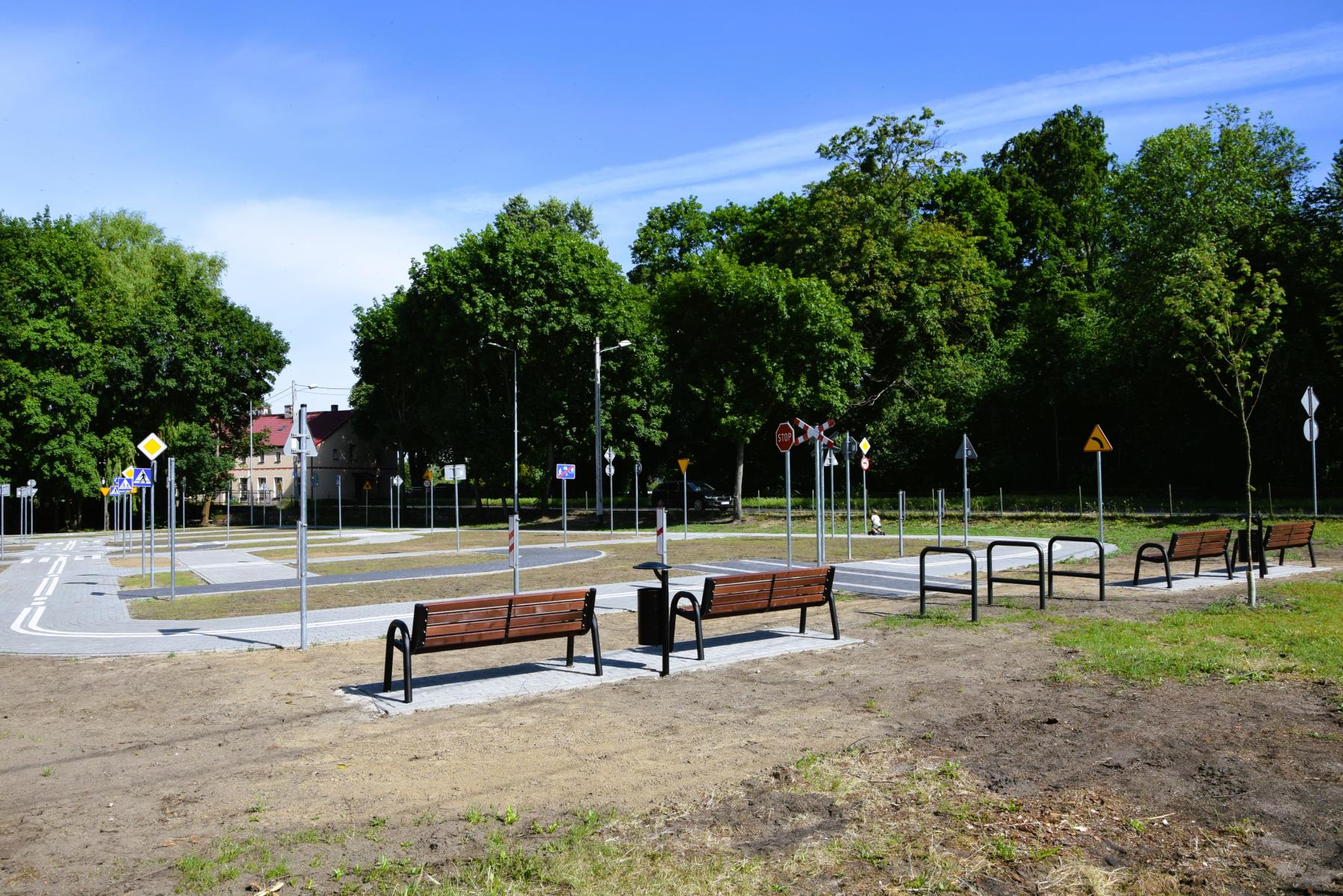 Plac miasteczka, z siecią dróg, znaki, ławeczki, stojaki na rowery. W tle duże zielone drzewa 