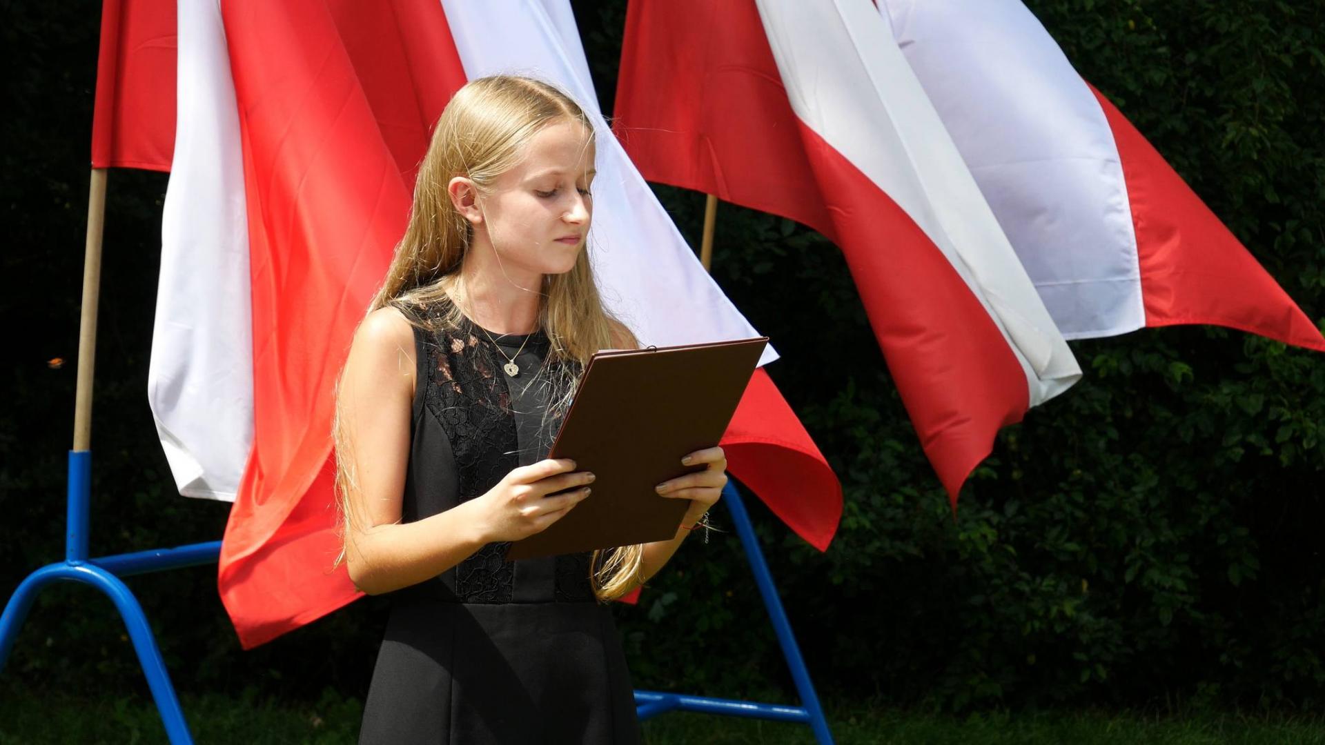 Młoda dziewczyna, w czarnej sukience, stoi, trzyma w rękach ciemną podkładkę. w tle powiewają biało-czerwone flagi. 