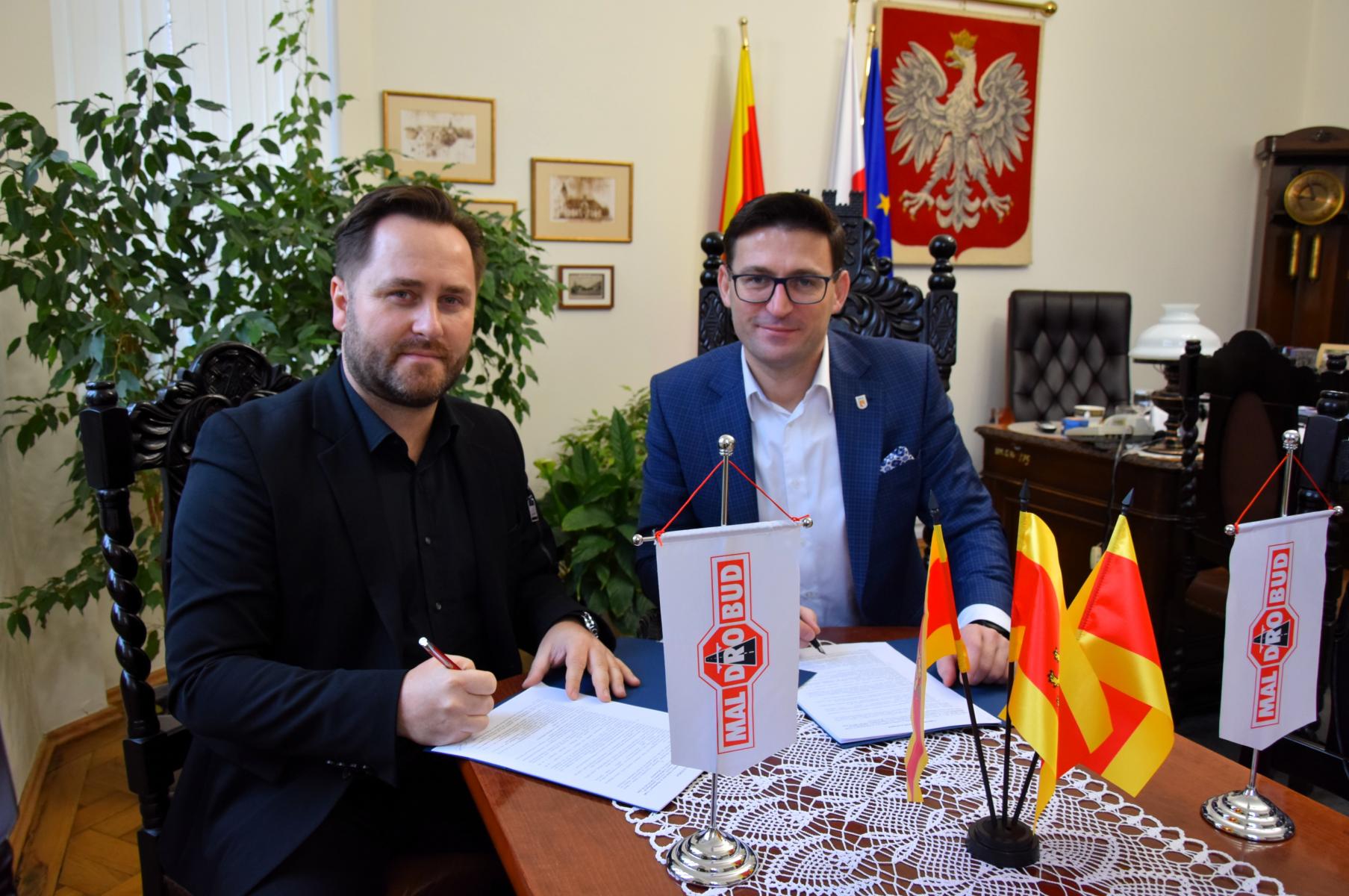 Od lewej siedzą przy stole i podpisują dokumenty: Łukasz Malinowicz, Grzegorz Kulbicki. Na stole stoją małe flagietki: białe z logiem maldrobud, żółto - czerwone flagi Dębna. W tle na ścianie wisi godło Polski. 