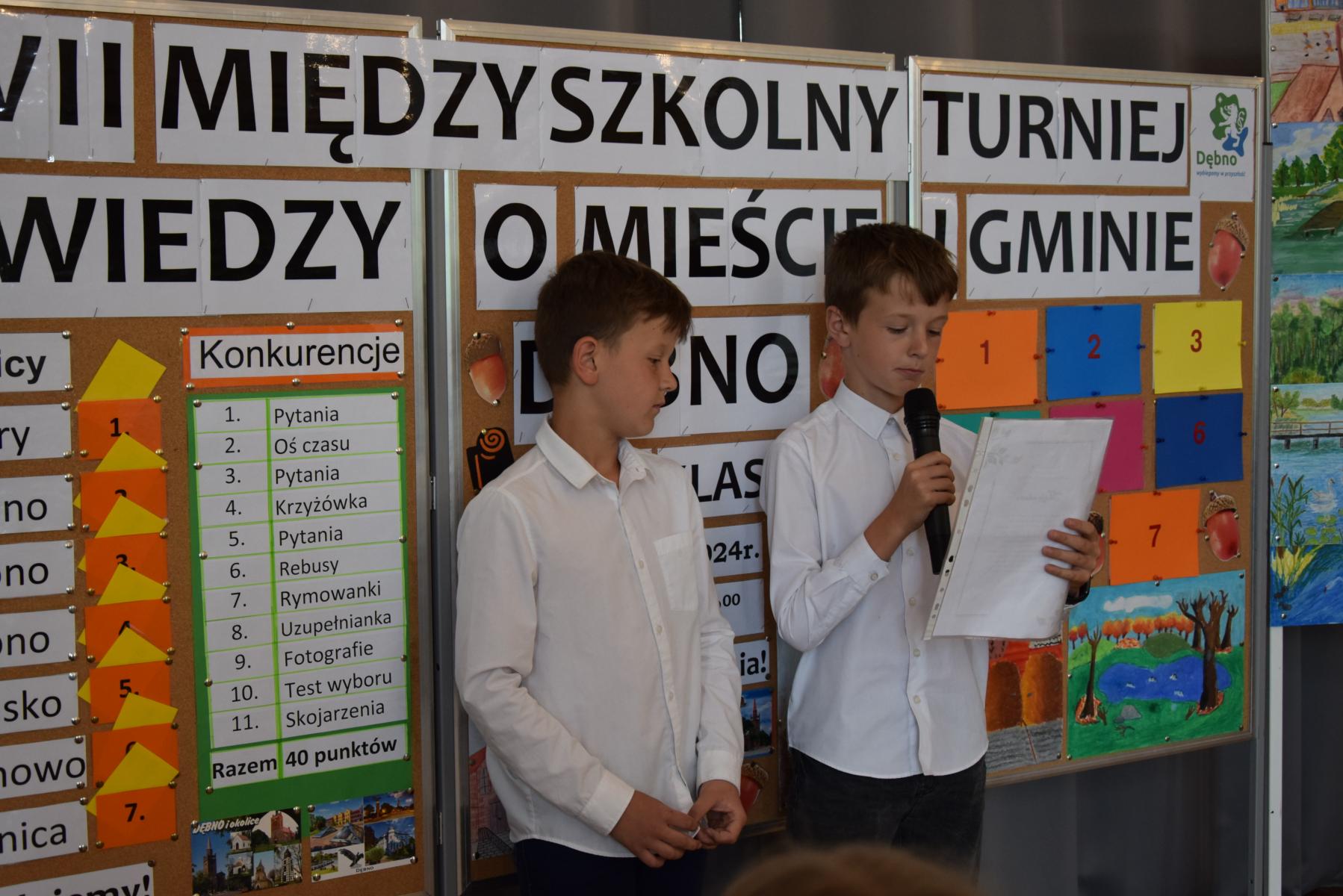 Dwóch uczniów w białych koszulach, jeden z nich trzyma mikrofon odczytuje wierszyk z kartki
