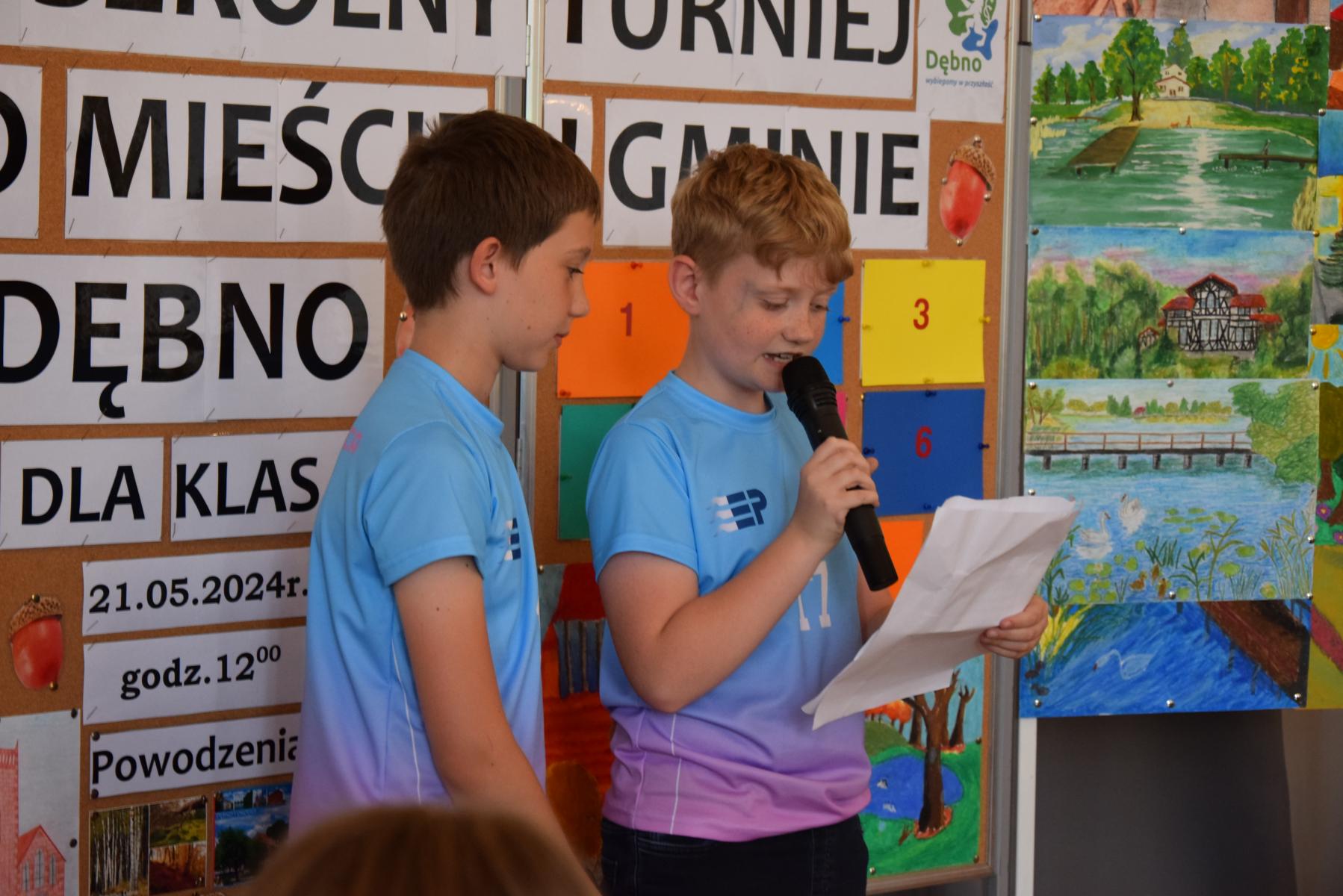 Dwóch chłopców w niebieskich koszulkach stoją. Jeden z nich trzyma mikrofon i czyta wierszyk z kartki