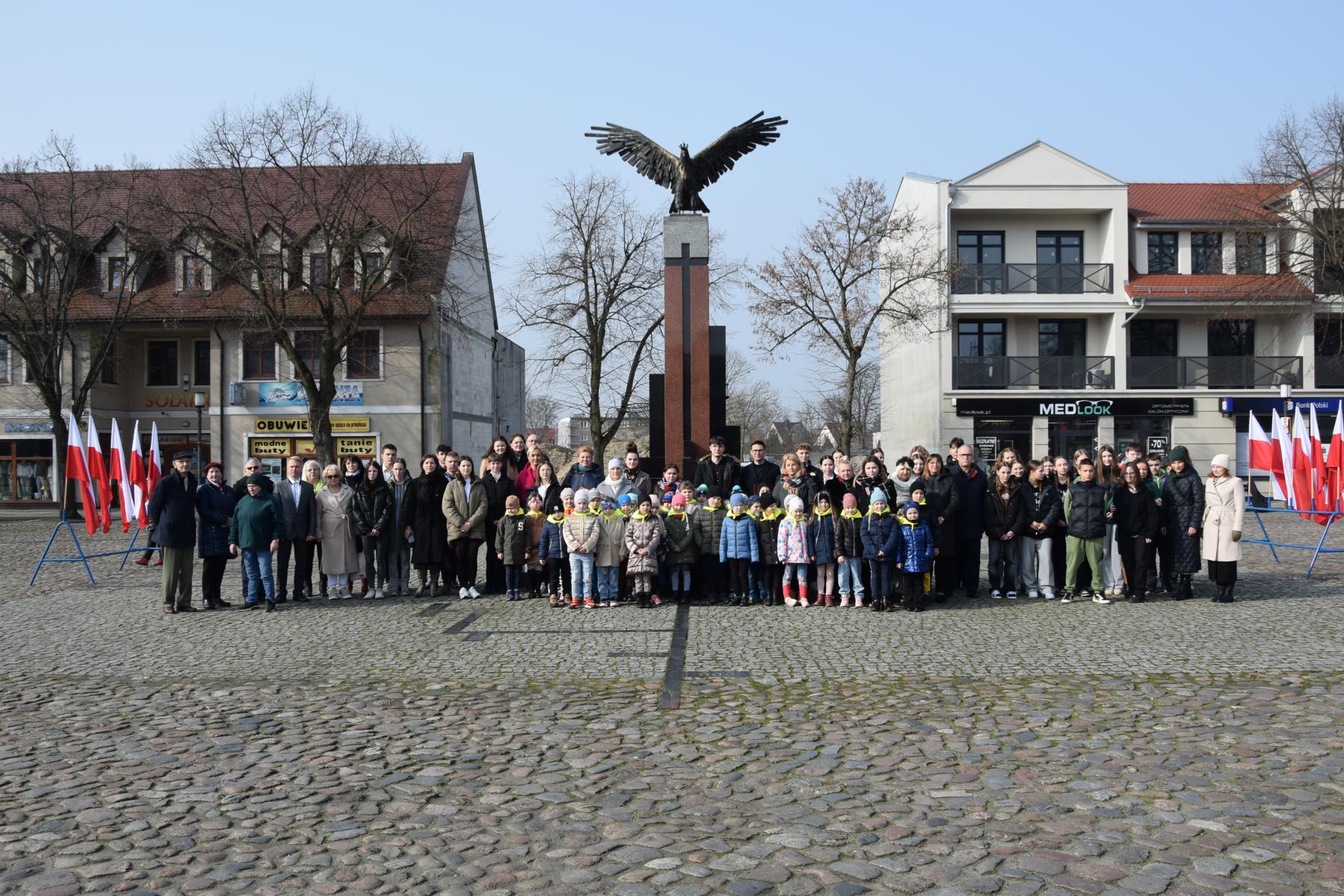 Grupa osób, dzieci, dorośli stoją przed pomnikiem Orła Białego