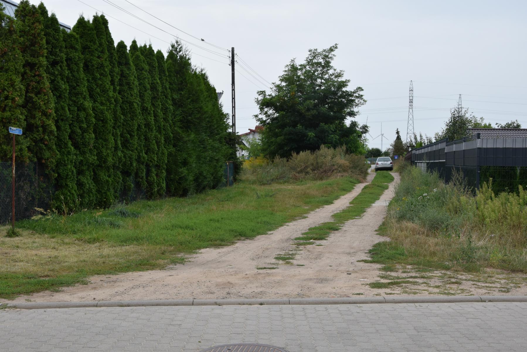 Obecny stan ulicy Łąkowej. Droga polna, po lewej stronie wysokie zielone tuje, po prawej ogrodzenie, w tle na drodze  samochód