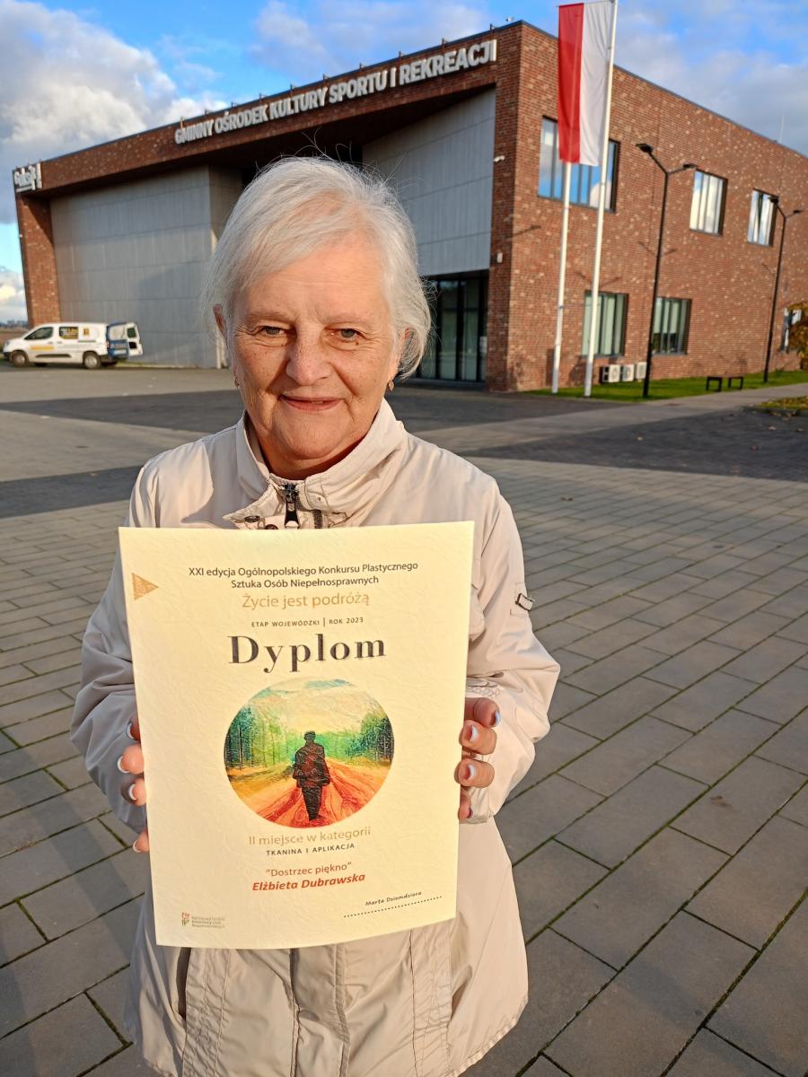 Starsza kobieta w rękach trzyma dyplom. W tle budynek, flaga Polski na maszcie, niebieskie niebo 