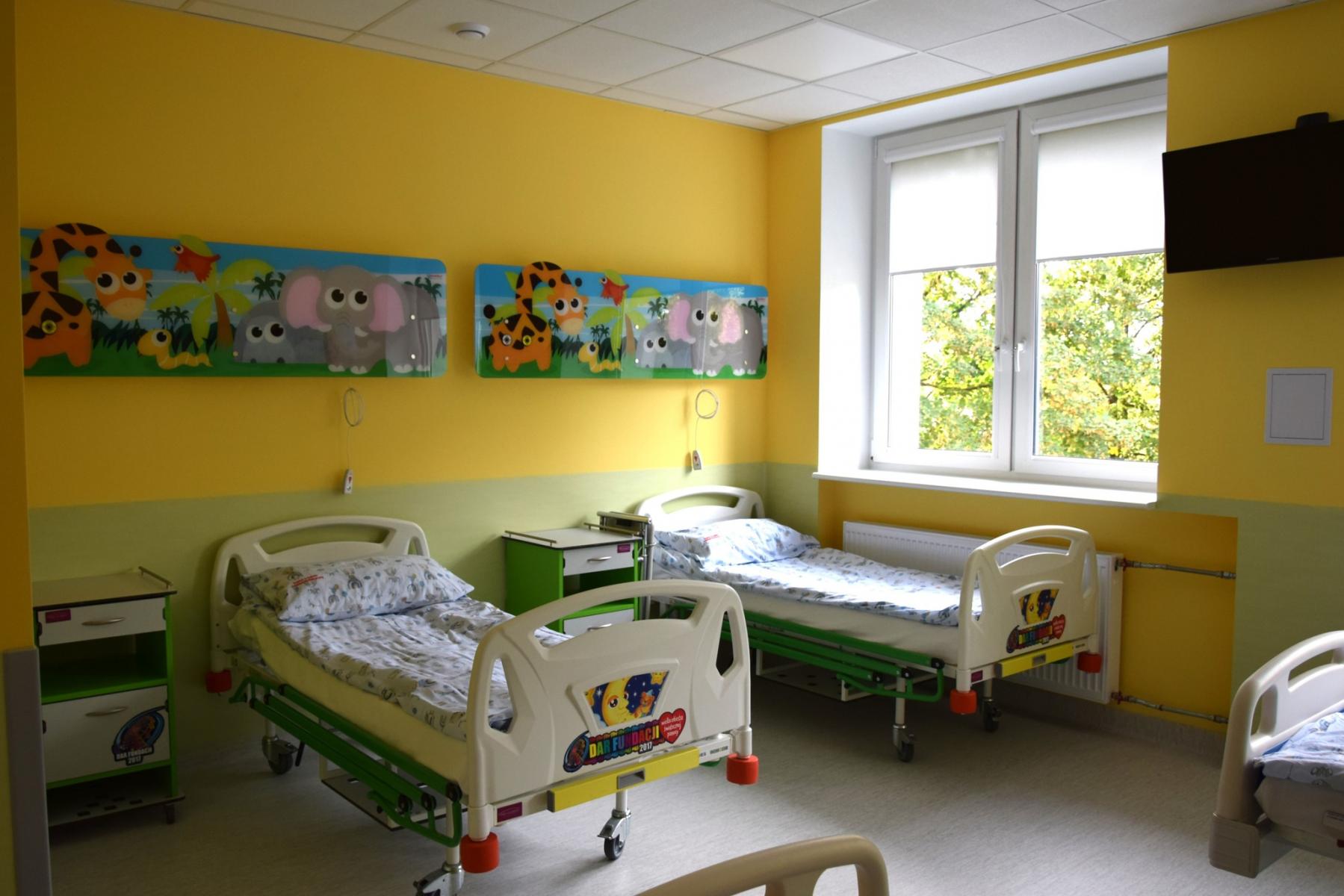 Żółto- zielona sala z kolorowymi obrazkami. Łóżka szpitalne i szafki obok