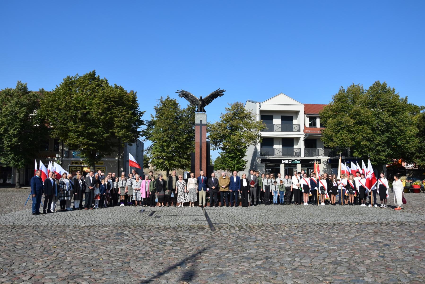 Powiększ zdjęcie. Duża grupa ludzi stoi, w tle pomnik Orła Białego, kamienica, drzewa, niebieskie niebo