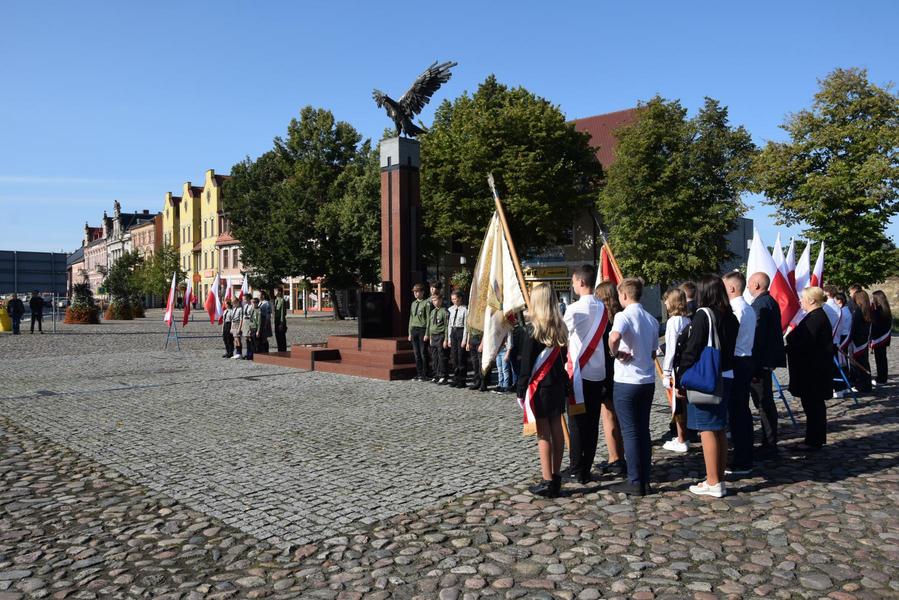 zgromadzeni na placu, uczniowie ze sztandarami, harcerze przy pomniku Orła Białego, w tle kamienice, drzewa
