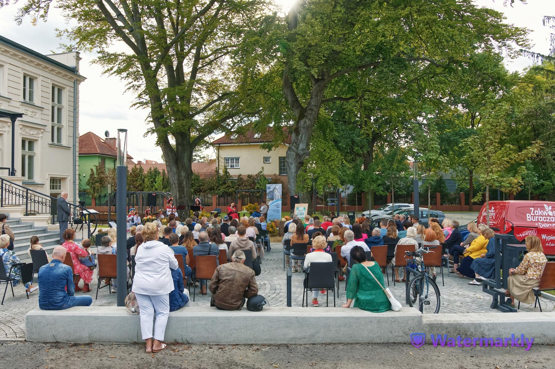 Zgromadzona publiczność siedzi na zewnątrz , w tle krzewy i drzewa. Po lewej stronie beżowy fragment willi ze schodami