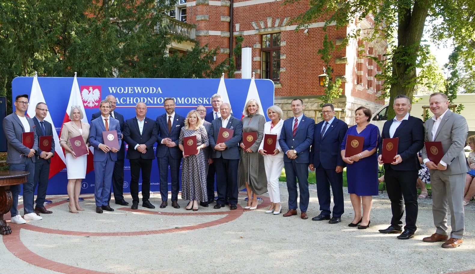 W rzędzie stoją mężczyźni i kobiety, w tle dwie flagi Polski, niebieska ścianka z napisem wojewoda zachodniopomorski, za nią drzewa i fragment budynku z czerwonej cegły
