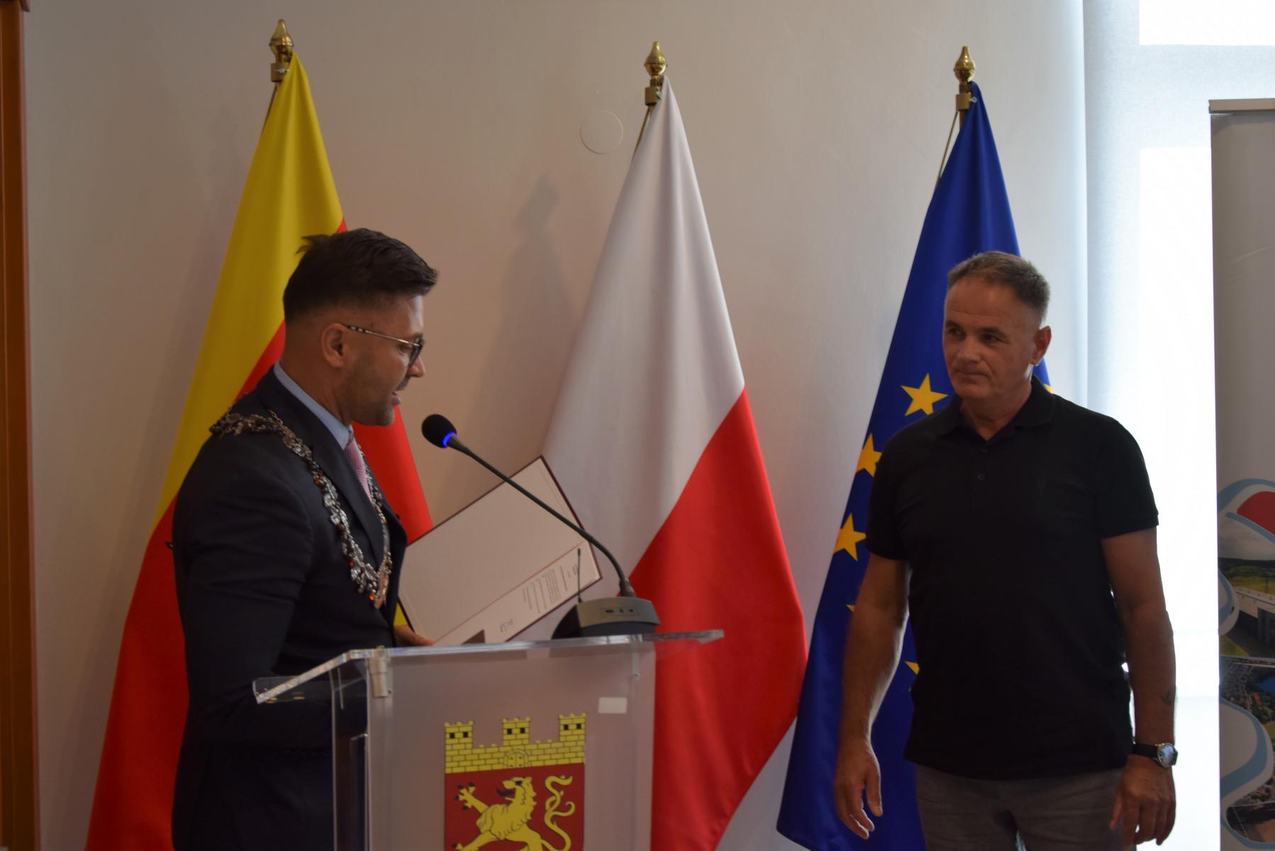 Od lewej przewodniczący przy mównicy, obok stoi mężczyzna Mieczysław Kukuruza. W tle flaga Dębna, Polski, UE