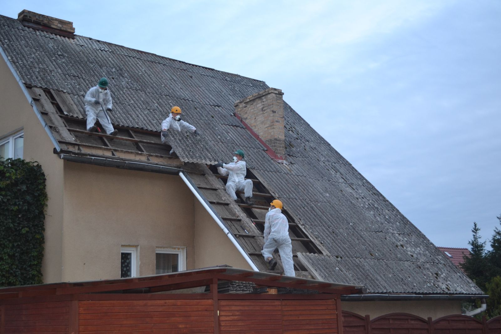 Pracownicy w białych kombinezonach usuwają płyty faliste z dachu