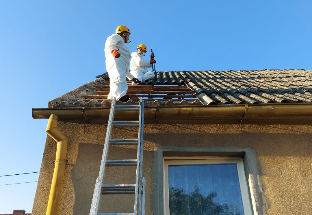 Pracownicy w kombinezonach na dachu usuwają płyty faliste z azbestem 